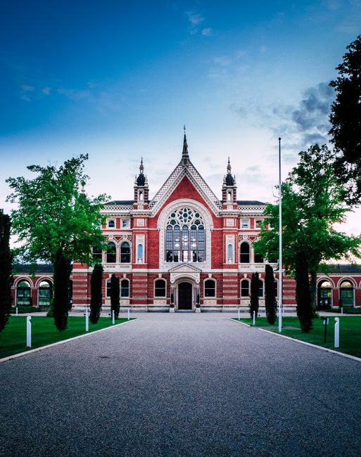 A large, historic red-brick building with intricate architectural details, including decorative arches, a prominent central stained-glass window, and multiple spires on the roof, situated in a landscaped area with a paved pathway leading up to its entrance. Flanking the building are two tall, leafy green trees, and the sky above is partly cloudy with a soft light, suggesting early evening or late afternoon. This image is related to house removals and relocation services by Removal Van Dulwich, showcasing the type of property where furniture transport and home relocation services may take place.