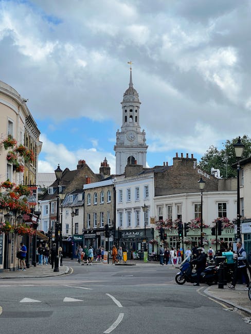 A busy street scene in Dulwich Village showing a mixture of historic and modern buildings, some with flower boxes on the windows. In the background, a tall white clock tower with a weather vane at the top rises above the surrounding structures. The street is populated with pedestrians strolling along the pavement, some carrying shopping bags, and others waiting at the crosswalk. Several vehicles, including a motorcycle and parked cars, are visible near the curb. On the left, buildings house various shops and cafes, with signage and outdoor displays. The weather appears partly cloudy with patches of blue sky peeking through. This urban environment, with its narrow street and diverse architecture, reflects the typical scene of a central Dulwich location, which Removal Van Dulwich regularly services for home relocations and furniture transport.