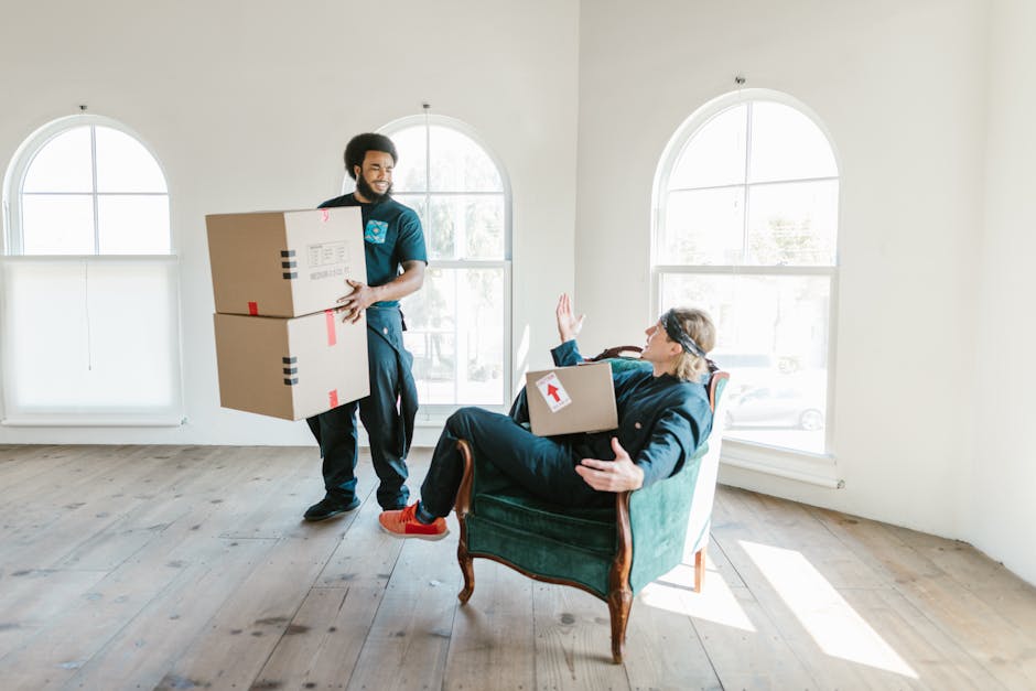 A man with dark curly hair and a beard, wearing a teal shirt, stands inside a bright room with white walls and large arched windows, preparing to move or load cardboard boxes for house relocation. He