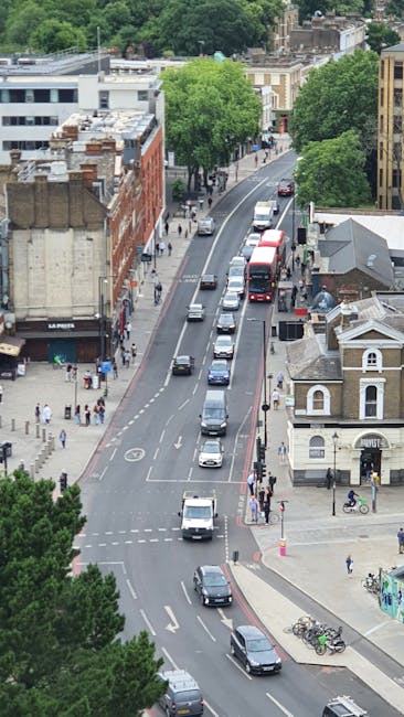 An aerial view of College Road in Dulwich showing a narrow, two-lane street with several vehicles, including a white van, a red bus, and multiple passenger cars, all queued in traffic. The street is lined with a mix of brick and stucco buildings, some with distinctive architectural features, and tall, leafy green trees providing shade along the sidewalks. Pedestrians are visible walking on the pavements, and bicycles are parked near a small white building on the corner. This scene captures an active residential area during daytime, illustrating typical urban traffic and street environment relevant for house removals and home relocation services, with visible space constraints that might complicate furniture transport and loading process, which Removal Van Dulwich handles with professional experience.